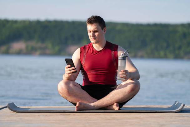 Genç bir adam telefona bakar ve nehir kıyısındaki yogadan sonra tekrar kullanılabilir bir şişeden su içer. Kilo verme ve doğada spor yapma kavramı.. - Fotoğraf, Görsel