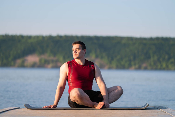 Młody człowiek ćwiczy jogę na brzegu rzeki. Koncepcja odchudzania i sportu w przyrodzie. - Zdjęcie, obraz