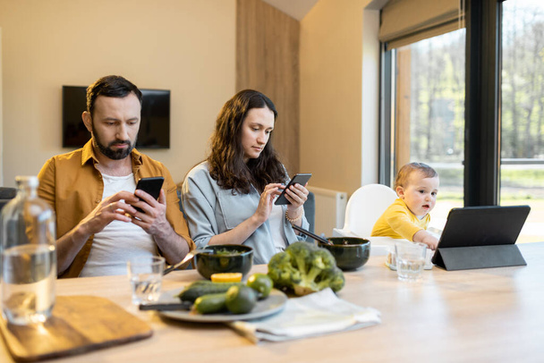 Pais e seu menino de um ano sentado em dispositivos móveis durante o almoço em casa - Foto, Imagem