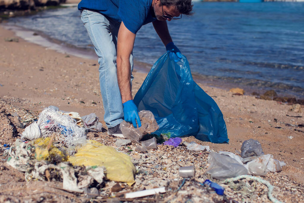 Чоловік доброволець у синій футболці з великою сумкою для збору сміття на пляжі. Концепція забруднення довкілля
 - Фото, зображення