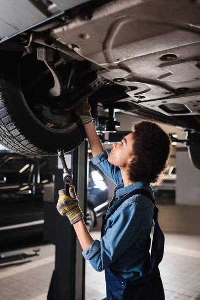 young african american mechanic in overalls holding wrench and repairing wheel of lifted car in garage  - 写真・画像