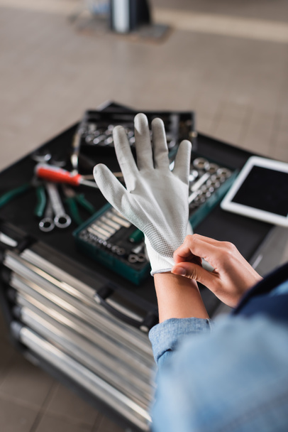 gedeeltelijke weergave van jonge monteur dragen handschoen bij de hand in de buurt gereedschapskist en digitale tablet in auto reparatie service - Foto, afbeelding