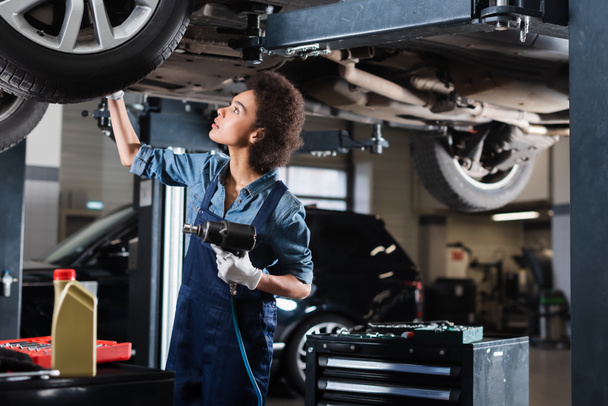 junger afrikanisch-amerikanischer Mechaniker in Overalls repariert angehobenes Auto mit Elektroschrauber in Garage - Foto, Bild