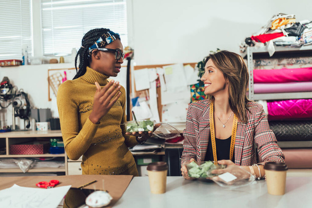 Deux jeunes femmes entrepreneures, ou créatrices de mode travaillant dans un atelier et faisant une courte pause déjeuner au travail - Photo, image