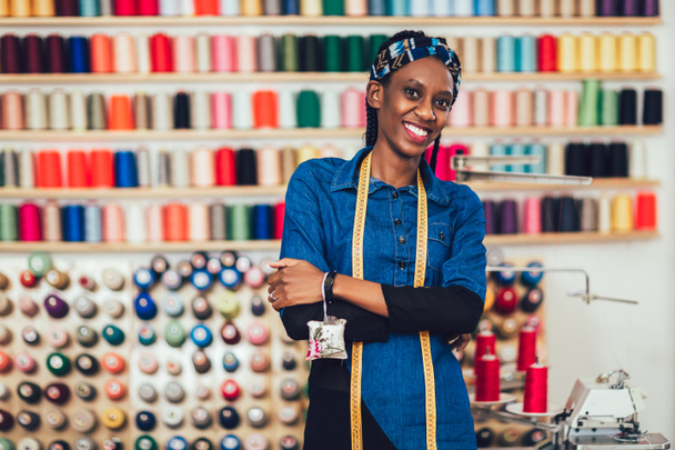 Πορτρέτο της χαρούμενης αφρικανίδας σχεδιάστριας μόδας στο στούντιο. Ιστορικό πολύχρωμου νήματος ραπτικής. - Φωτογραφία, εικόνα