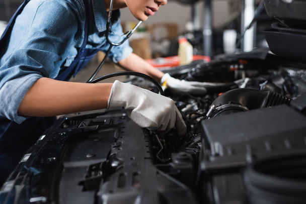 partial view of young african american mechanic in gloves and stethoscope working with car motor in garage - Photo, Image