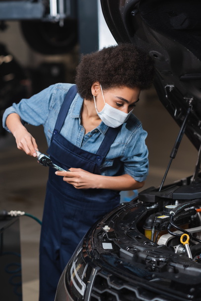 focused young african american mechanic in protective mask holding wrench and working with car motor in garage - Foto, Imagen