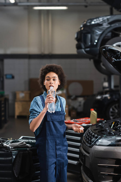 junger afrikanisch-amerikanischer Mechaniker trinkt Wasser aus Flasche neben Auto in Garage - Foto, Bild