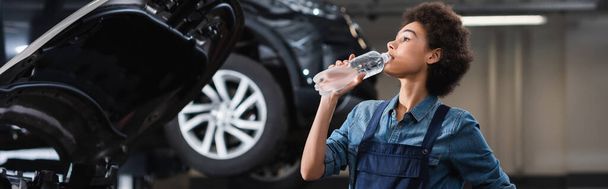 mladý africký americký mechanik v montérkách pitná voda z láhve u auta v garáži, banner - Fotografie, Obrázek