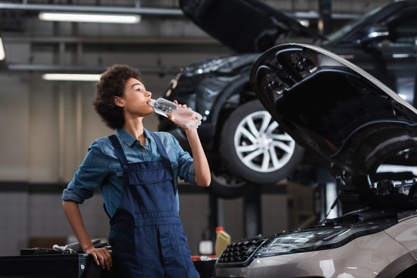 junger afrikanisch-amerikanischer Mechaniker in Overalls trinkt Wasser aus Plastikflasche neben Auto in Garage - Foto, Bild
