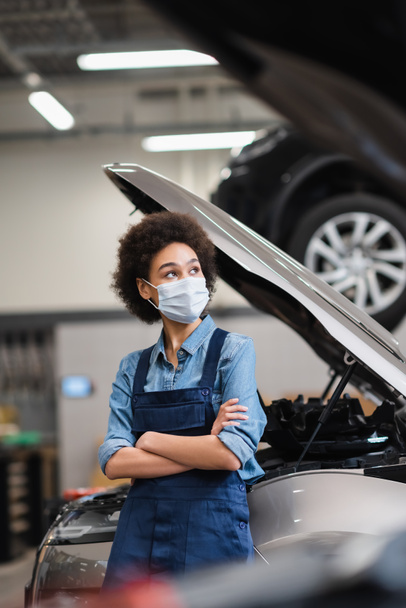 young african american mechanic in protective mask standing with crossed arms near car with open hood in garage - Photo, Image