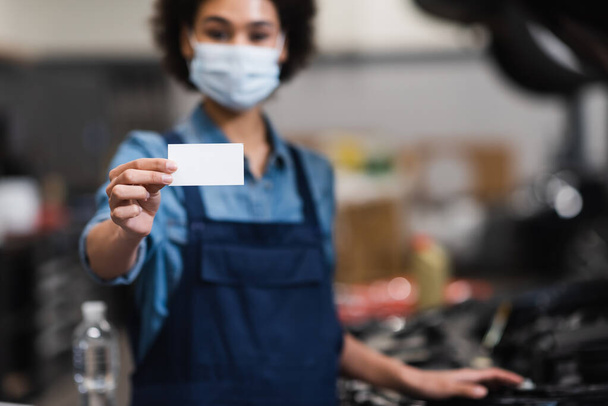 empty card in hand of blurred young african american mechanic in protective mask in garage - 写真・画像