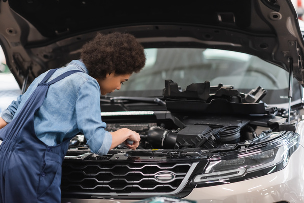 young african american mechanic working with motor of car with open hood in garage - Foto, Bild