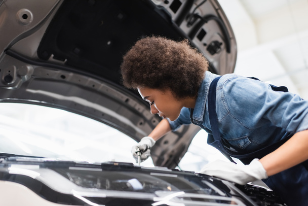 молодой африканский американский механик фиксирующий мотор в машине с открытым капотом в гараже - Фото, изображение