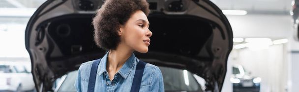 giovane meccanico afroamericano in piedi vicino al cappuccio aperto e guardando altrove nel servizio di riparazione auto, banner - Foto, immagini