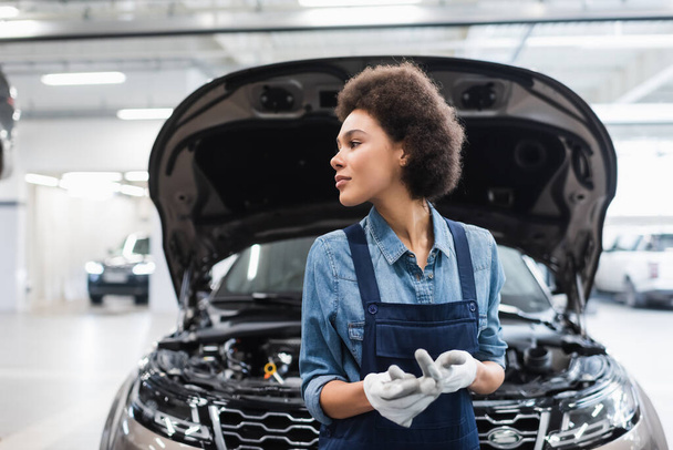 jeune mécanicien afro-américain debout près de la voiture avec capot ouvert et regardant loin dans le service de réparation automobile - Photo, image