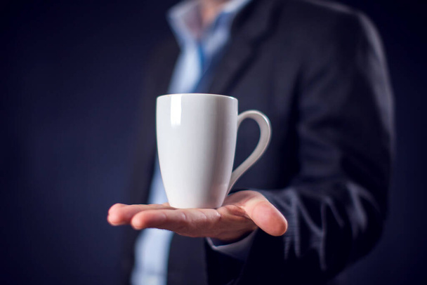 бизнесмен в костюме держит чашку чая или кофе в руке на фоне черного фона. Концепция кофе-брейка - Фото, изображение