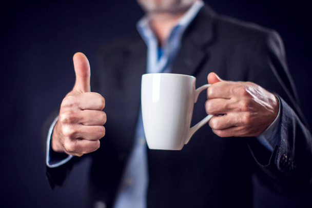 бизнесмен в костюме держит чашку чая или кофе в руке на фоне черного фона. Концепция кофе-брейка - Фото, изображение