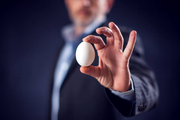 Geschäftsmann im Anzug mit einem weißen Ei in der Hand vor schwarzem Hintergrund - Foto, Bild
