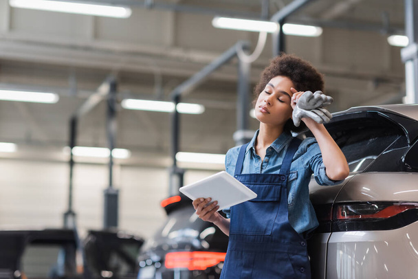 Müder junger afrikanisch-amerikanischer Mechaniker mit geschlossenen Augen, der mit digitalem Tablet in der Garage steht und die Hand am Kopf hält - Foto, Bild
