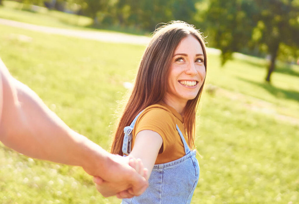 Jong gelukkig stel loopt samen in een park en hand in hand - Vriendin kijkt terug op haar vriend en glimlacht naar hem - Vrienden hebben plezier samen - Outdoor recreatie lifestyle concept - Foto, afbeelding