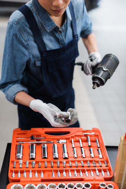partial view of young mechanic with hands in gloves holding electric screwdriver near toolbox in auto repair service - Photo, Image