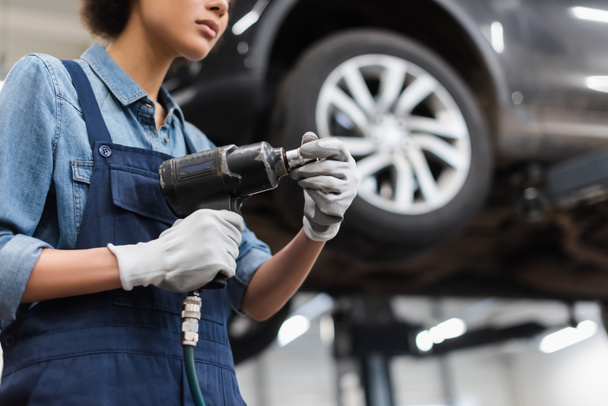 vista parcial del joven mecánico con las manos en guantes sosteniendo destornillador eléctrico en servicio de reparación de automóviles - Foto, imagen