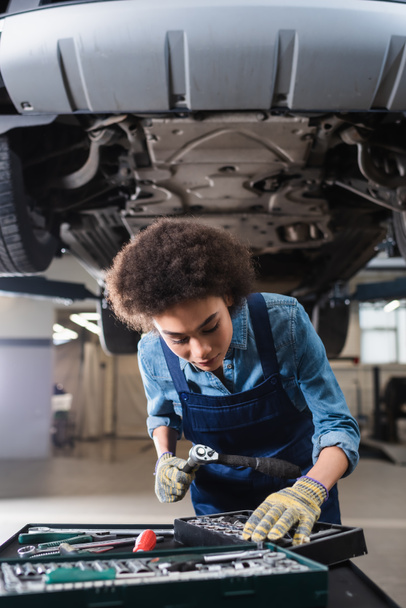 young african american mechanic standing beneath lifted car in garage - Foto, Imagen