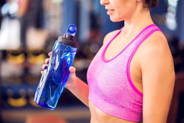 Женщина в розовом топе держит голубую бутылку воды, отдыхая в тренажерном зале. люди, фитнес и здравоохранение концепции - Фото, изображение