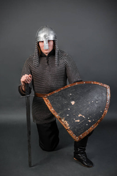 Portrait of a medieval warrior of the late viking era and the beginning of the crusades sitting on his knee. Knight in chain mail and helmet armed with shield and sword isolated on dark background. - Photo, image