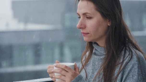 Mulher caucasiana fica na varanda durante a queda de neve com xícara de café quente ou chá. Ela olha para os flocos de neve e respira no ar fresco gelado - Filmagem, Vídeo