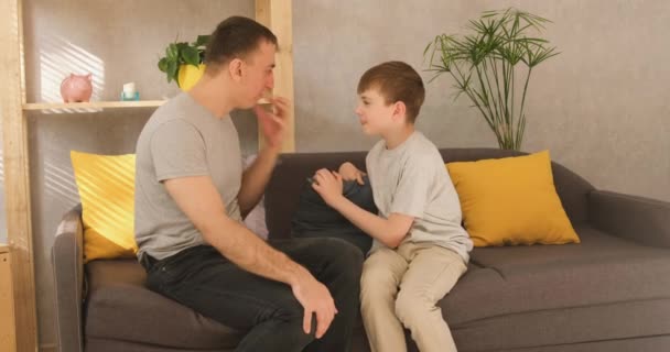 Ojciec emocjonalnie komunikuje się ze swoim młodym synem siedzącym na kanapie w salonie. Widok z przodu. światło dzienne - Materiał filmowy, wideo