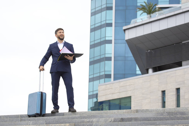 glücklicher Geschäftsmann steht mit Reisekoffer auf den Stufen vor dem Hintergrund eines Bürogebäudes, Geschäftsunterlagen in der Hand - Foto, Bild