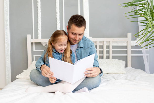 ο πατέρας διαβάζει ένα βιβλίο ή σπουδάζει με το παιδί της κόρης του πριν πάει για ύπνο ή διασκεδάζει - Φωτογραφία, εικόνα