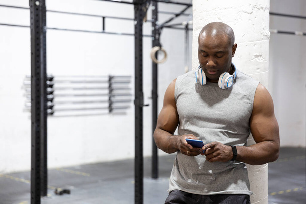 Silny czarny mężczyzna korzystający z telefonu komórkowego w siłowni. Robi sobie przerwę. Miejsce na tekst. - Zdjęcie, obraz