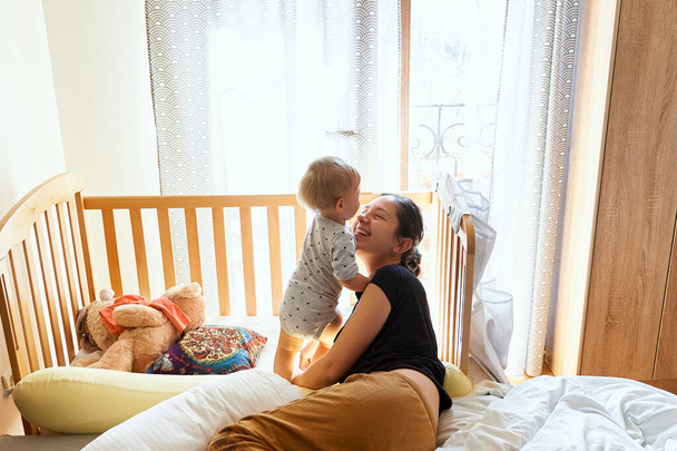 Smiling mom lies on the bed near the standing baby on the background of a crib and toys - Фото, изображение