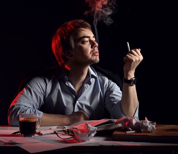 Adult man in casual clothing sits behind desk, writing table with book and papers, drinks tea, coffee and smokes - Foto, afbeelding