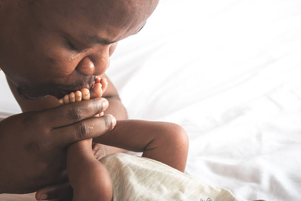 Imagens de perto de um pai afro-americano beijando o pé, seu bebê recém-nascido, está deitado na cama branca, com amor e feliz, conceito para a família afro-americana e bebê recém-nascido de pele preta - Foto, Imagem