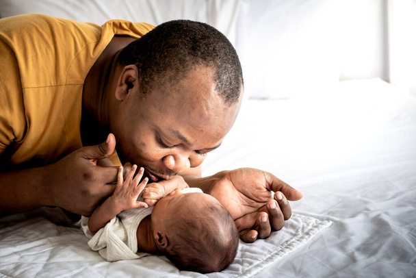 An African American father kissing hand, his 12-day-old baby newborn son lying in bed in a white bedroom, with happy, concept to African American family and newborn - Foto, Bild