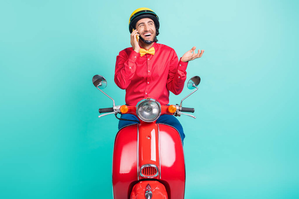 Photo of optimistic brunette guy ride on bike talk telephone wear red shirt helmet isolated on teal color background - Foto, immagini