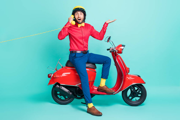 Full size photo of young shocked amazed man sit moped talking on telephone isolated on turquoise color background - 写真・画像