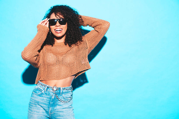 Beautiful black woman with afro curls hairstyle.Smiling model in trendy summer clothes. Sexy carefree female posing near blue wall in studio. Tanned and cheerful in sunglasses - Foto, imagen
