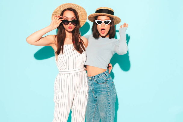 Two young beautiful smiling hipster female in trendy summer white t-shirt and jeans clothes.Sexy carefree women posing near light blue wall in studio.Positive and cheerful models in hats - Foto, Imagem