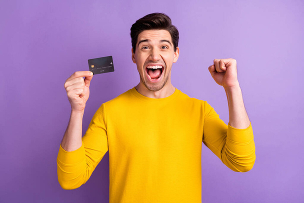 Portrait of astonished guy fist up celebrate open mouth shout yes hold debit card isolated on purple color background - Foto, imagen