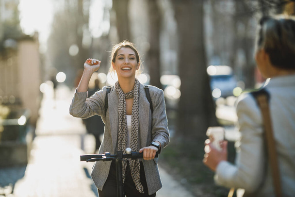 Uma jovem empresária montando uma scooter elétrica e cumprimenta alguém em seu caminho para o trabalho. - Foto, Imagem