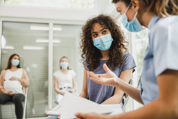 Upset African American teenage girl with protective mask talks with nurse while waits for gynecologist check up. - Photo, Image