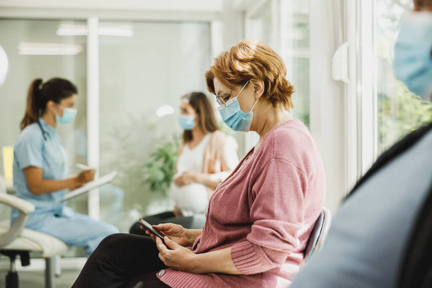 Senior vrouw met een gezichtsmasker zittend in een wachtkamer van een ziekenhuis en surfen op haar smartphone. - Foto, afbeelding
