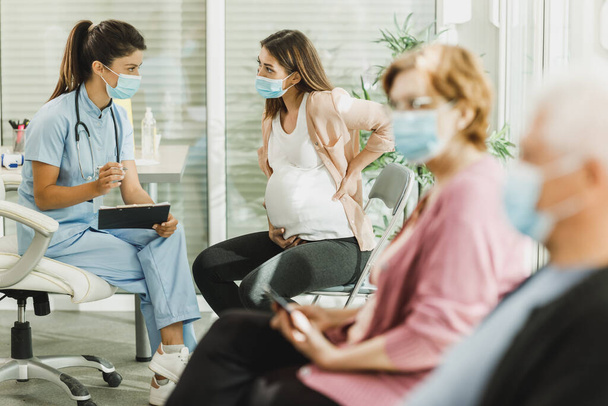 Nurse talking to young pregnant woman and taking notes before covid-19 vaccine at hospital waiting room. - Φωτογραφία, εικόνα