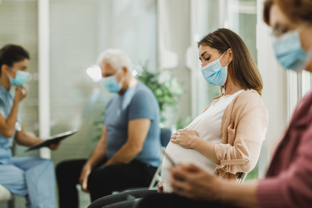 Молодая беременная женщина в маске сидит в приемной и ждет медицинского осмотра во время пандемии коронного вируса. - Фото, изображение