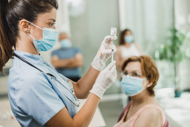 Hemşire elinde şırınga tutuyor ve maske takan yaşlı bir kadının omzuna Covid-19 aşısı yapıyor.. - Fotoğraf, Görsel
